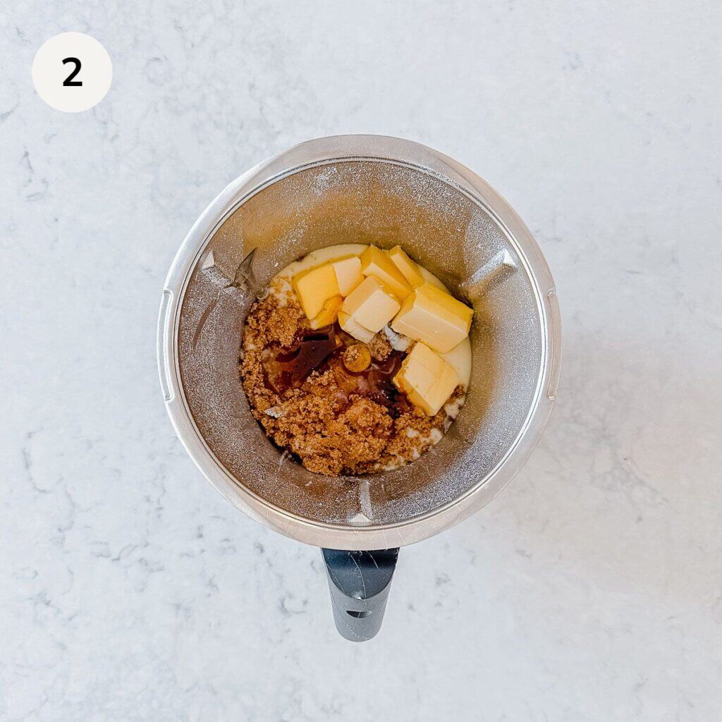 A mixing bowl with cubes of butter, brown sugar, and liquid vanilla on top of the ingredients, viewed from above, on a white marble surface. The number 2 is in a white circle in the top left corner.