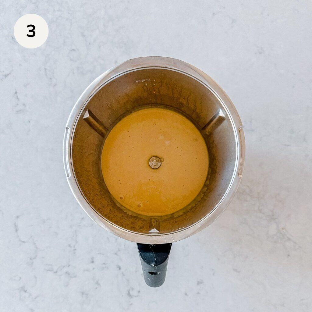 A top-down view of a blender holding a creamy, light brown liquid mixture. The countertop is a light, speckled surface, and a white circle with the number 3 is in the top left corner.