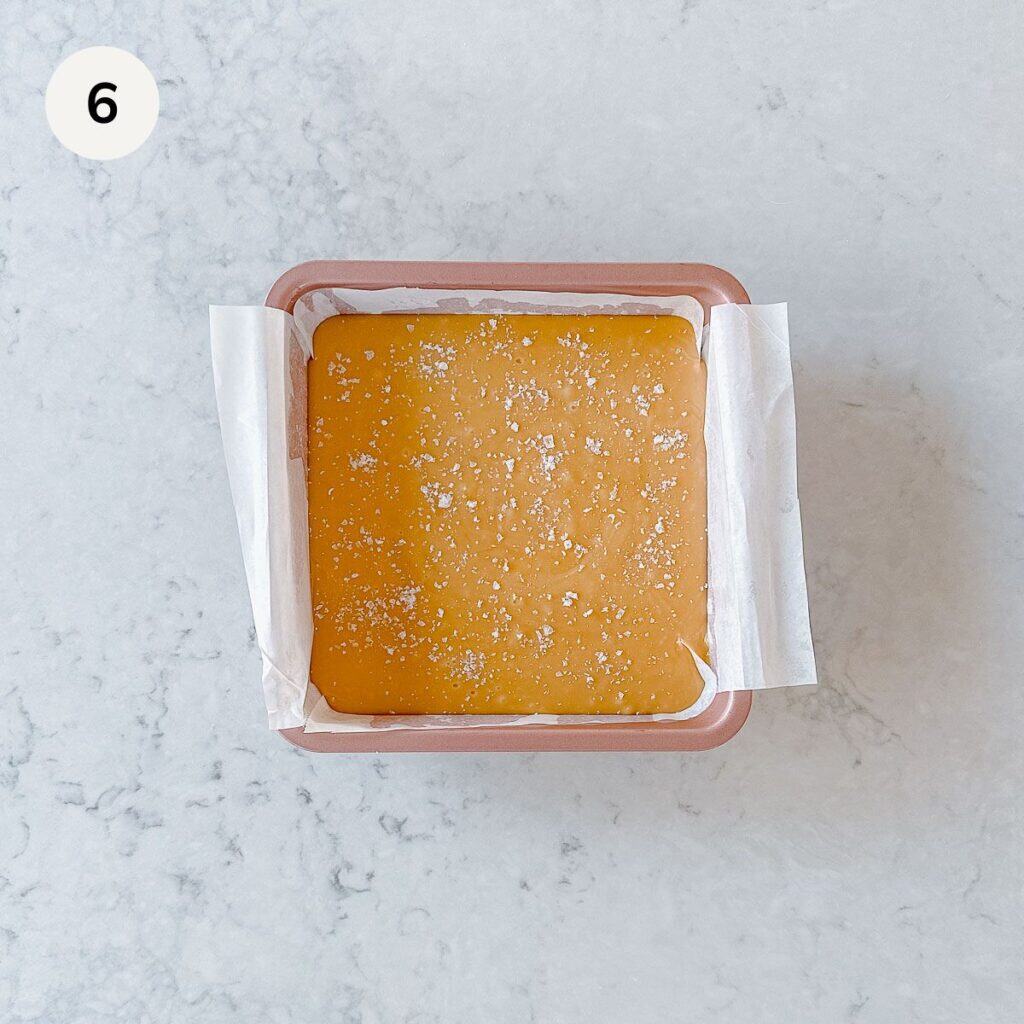 A square baking dish lined with parchment paper holds a slab of golden caramel sprinkled with sea salt, viewed from above on a light marble surface. A number 6 is in the top left corner.