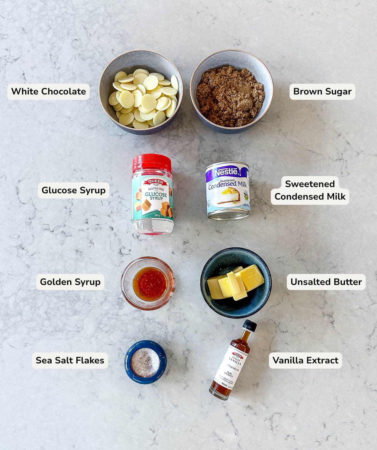 Ingredients for fudge arranged on a countertop: white chocolate chips, brown sugar, glucose syrup, sweetened condensed milk, golden syrup, unsalted butter, sea salt flakes, and vanilla extract, each labeled.
