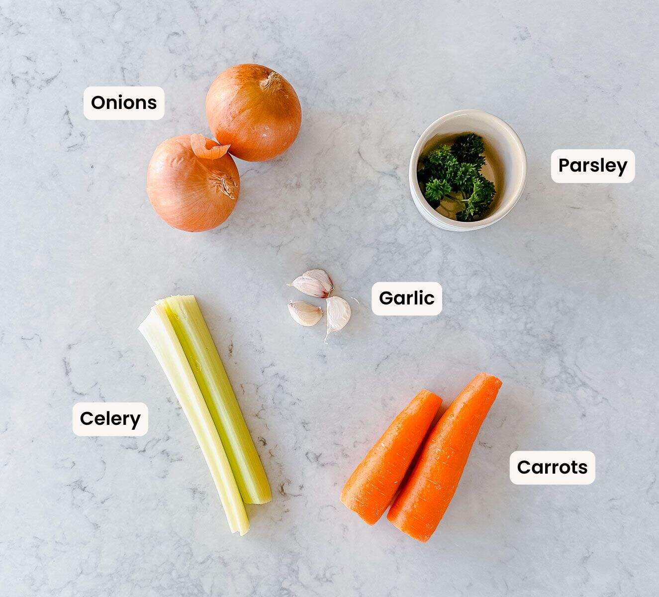 Onions, carrots, celery, parsley, garlic and parsley on a marble benchtop which are labelled.