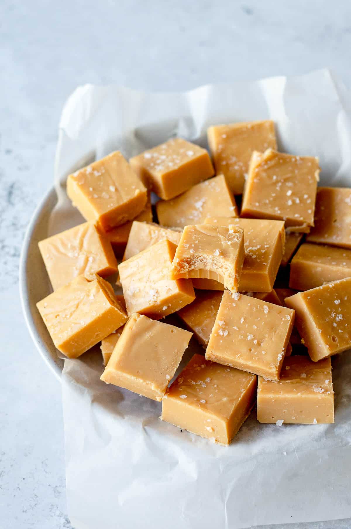 A plate lined with parchment paper holds several pieces of light brown caramel fudge, each sprinkled with coarse sea salt. The fudge squares are neatly arranged and have a smooth, creamy texture.