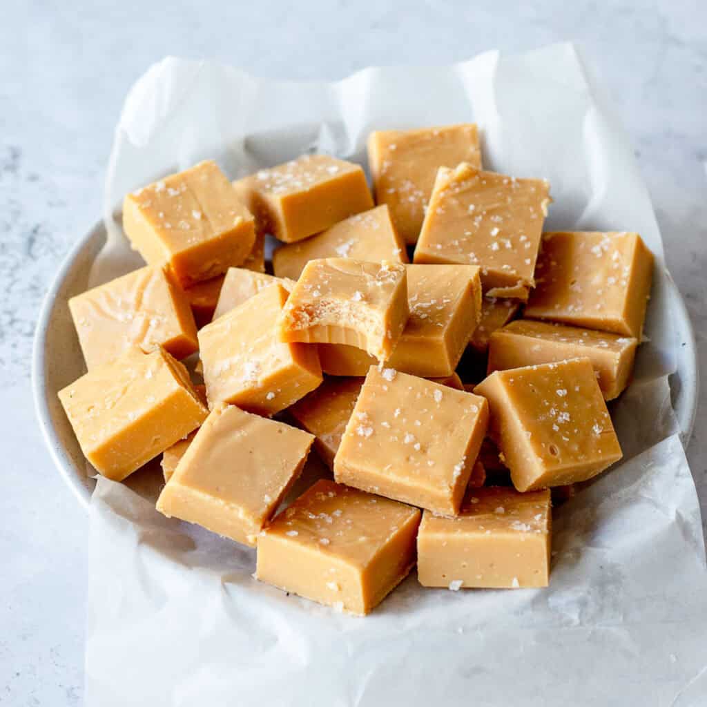 A plate lined with baking paper, holds several squares of creamy caramel fudge, topped with a sprinkle of coarse sea salt. One piece near the center has a bite taken out of it.