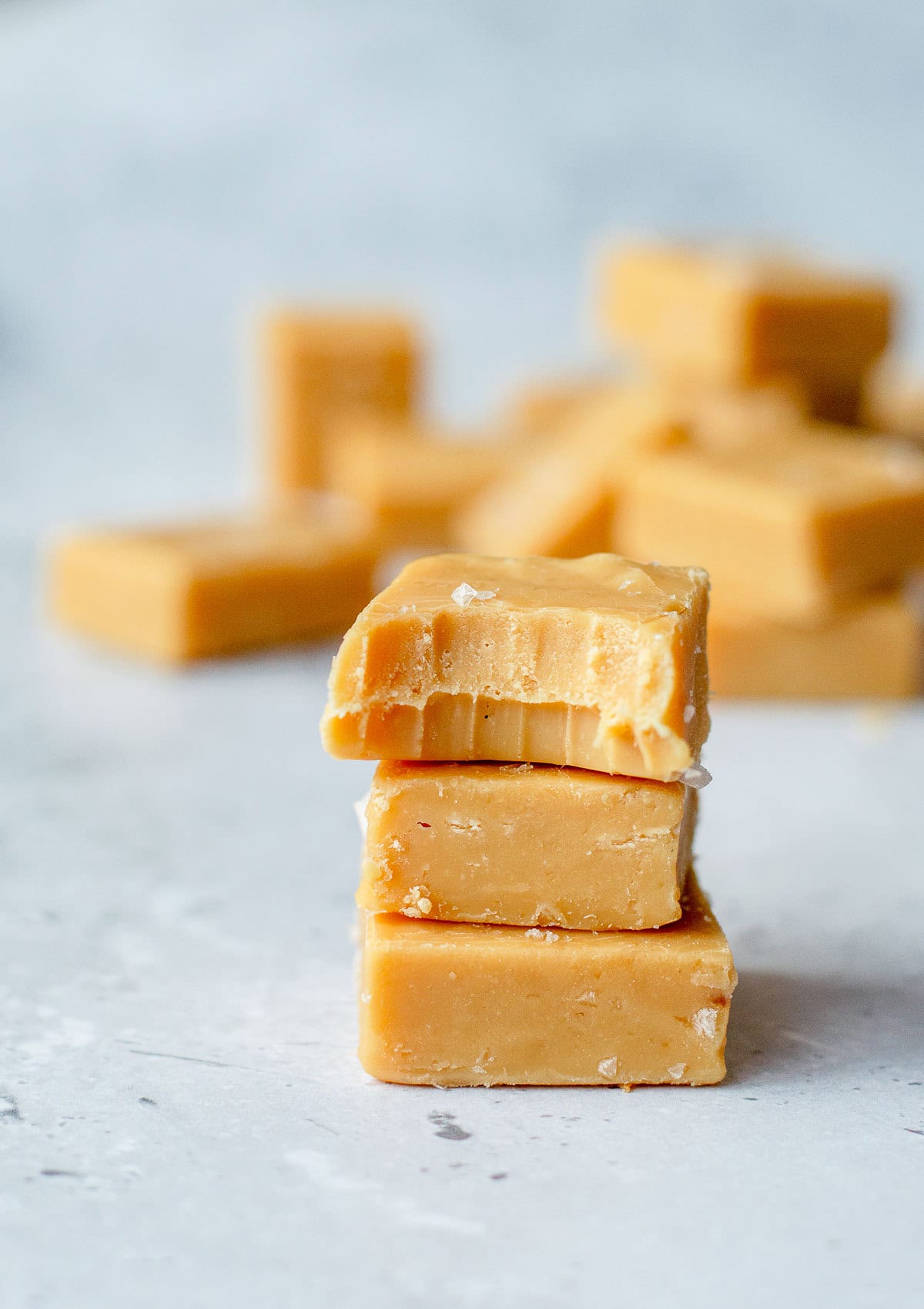 A plate lined with parchment paper holds several pieces of light brown salted caramel fudge, with one piece showing a bite taken out of it. The fudge is topped with flaky sea salt.