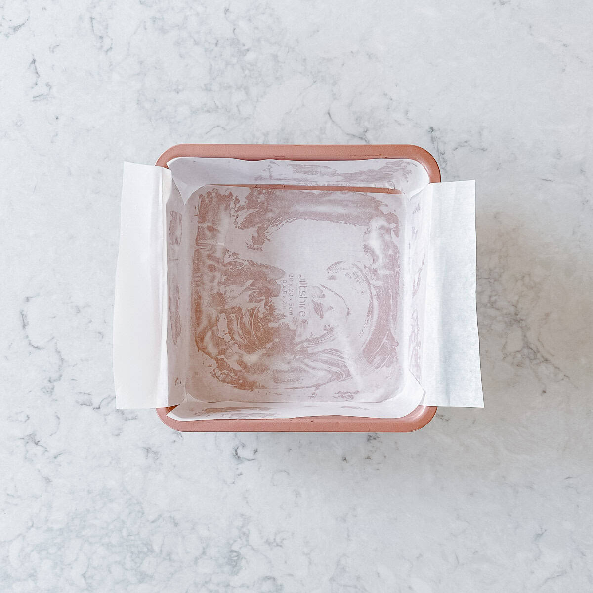 A square baking tin lined with baking paper, on a light grey marble benchtop.