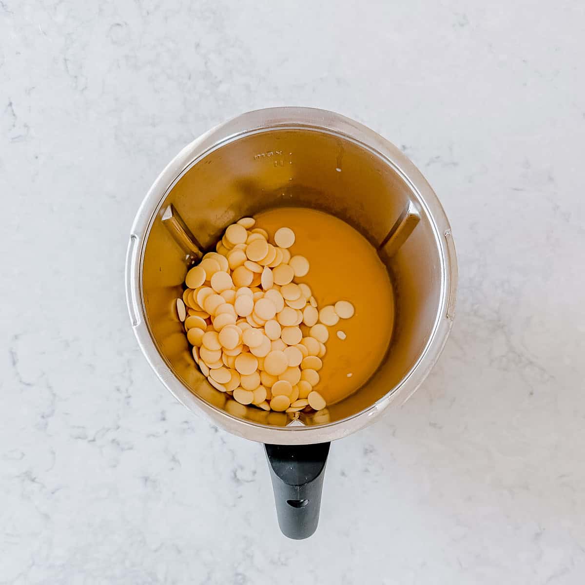 An overhead image of white chocolate melts being added to fudge mixture in a Thermomix Bowl.