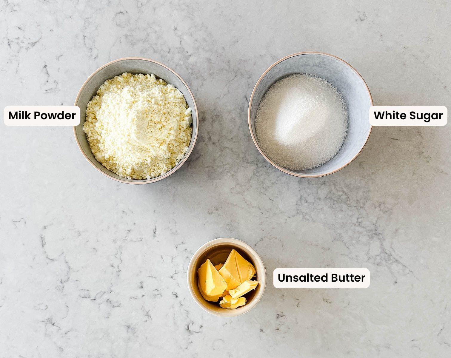 Three bowls on a marble surface: one with milk powder, one with white sugar, and a smaller bowl with pieces of unsalted butter. Each bowl is labeled with its ingredient name.
