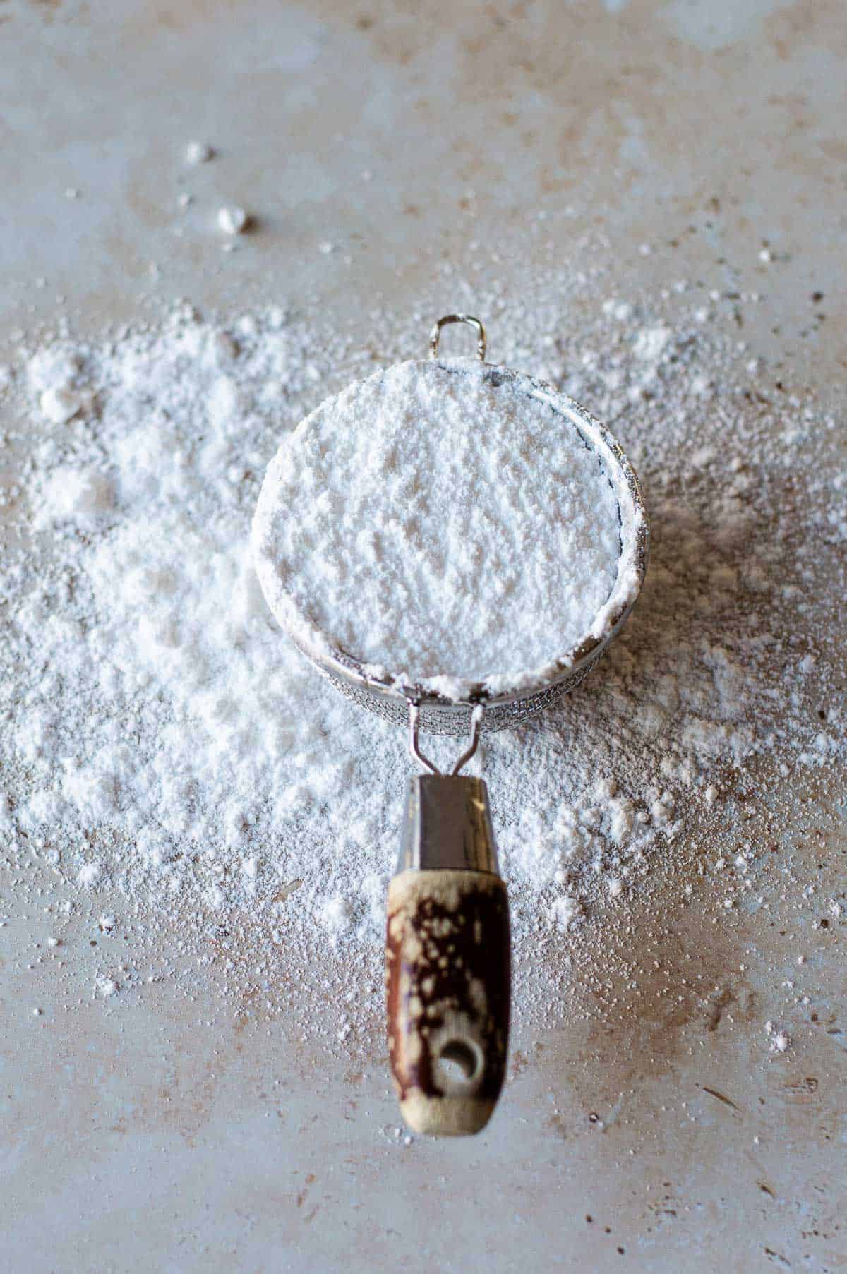 Homemade Icing Sugar in a small sieve, overflowing on a grey benchtop.
