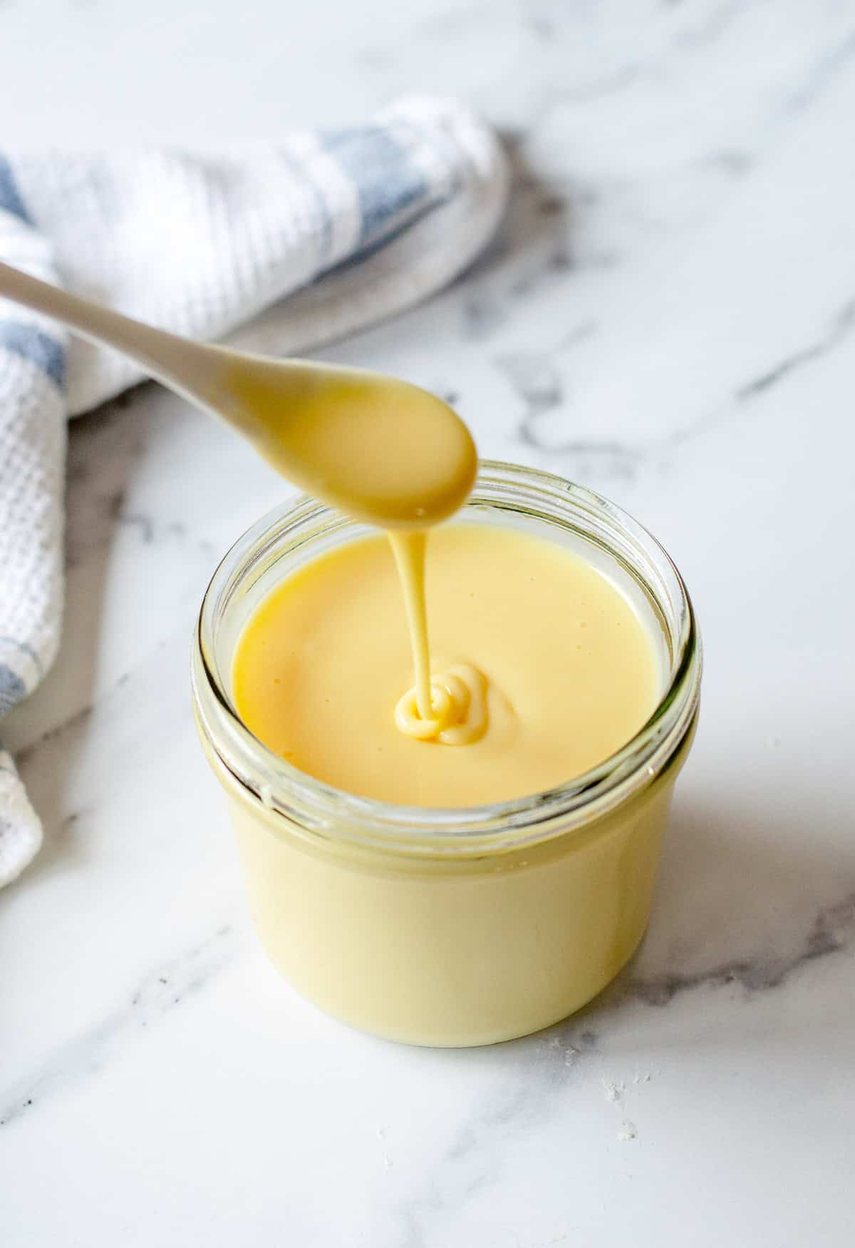 Thick, creamy condensed milk being drizzled from a spoon into a glass jar on a white marble bench, with a white and blue tea towel in the background.