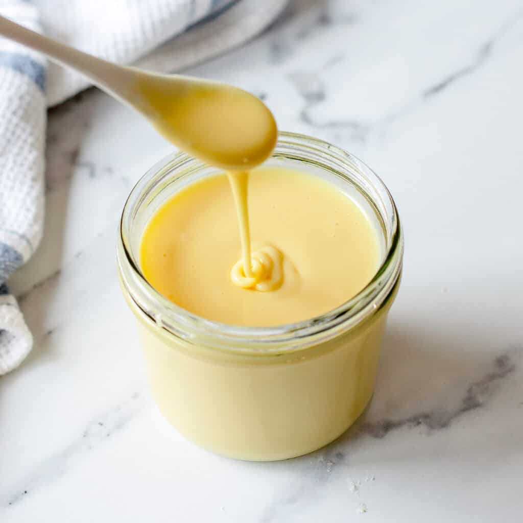 A spoon drizzling thick, creamy condensed milk into a glass jar on a white marble surface, with a white and blue towel in the background.