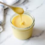 A spoon drizzling thick, creamy condensed milk into a glass jar on a white marble surface, with a white and blue towel in the background.