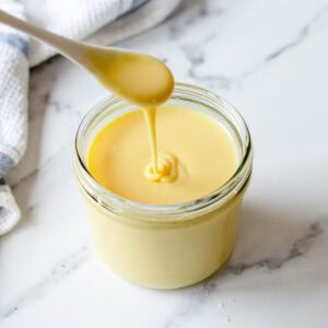 A spoon drizzling thick, creamy condensed milk into a glass jar on a white marble surface, with a white and blue towel in the background.