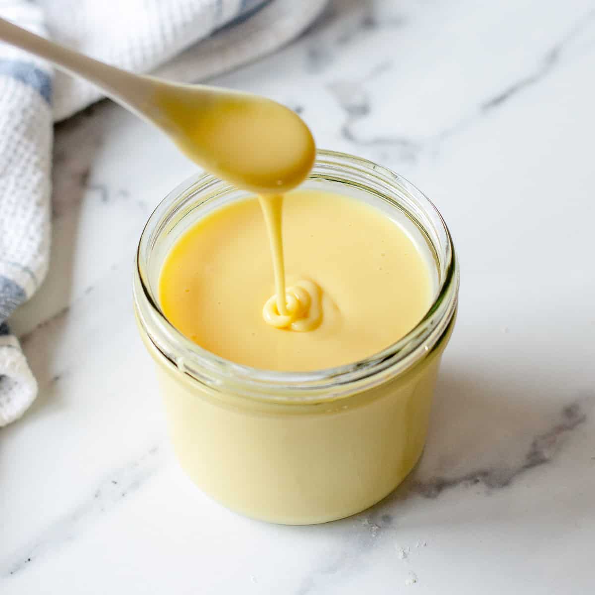 A spoon drizzling thick, creamy condensed milk into a glass jar on a white marble surface, with a white and blue towel in the background.