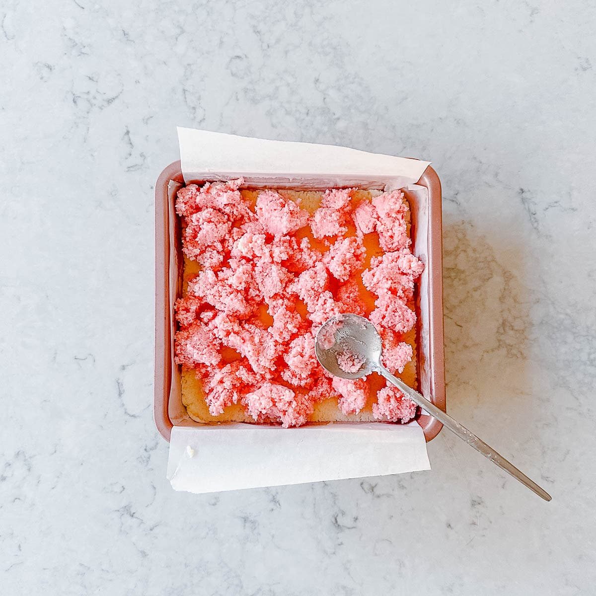 Spoonfuls of pink coconut ice in a pink square baking tin.