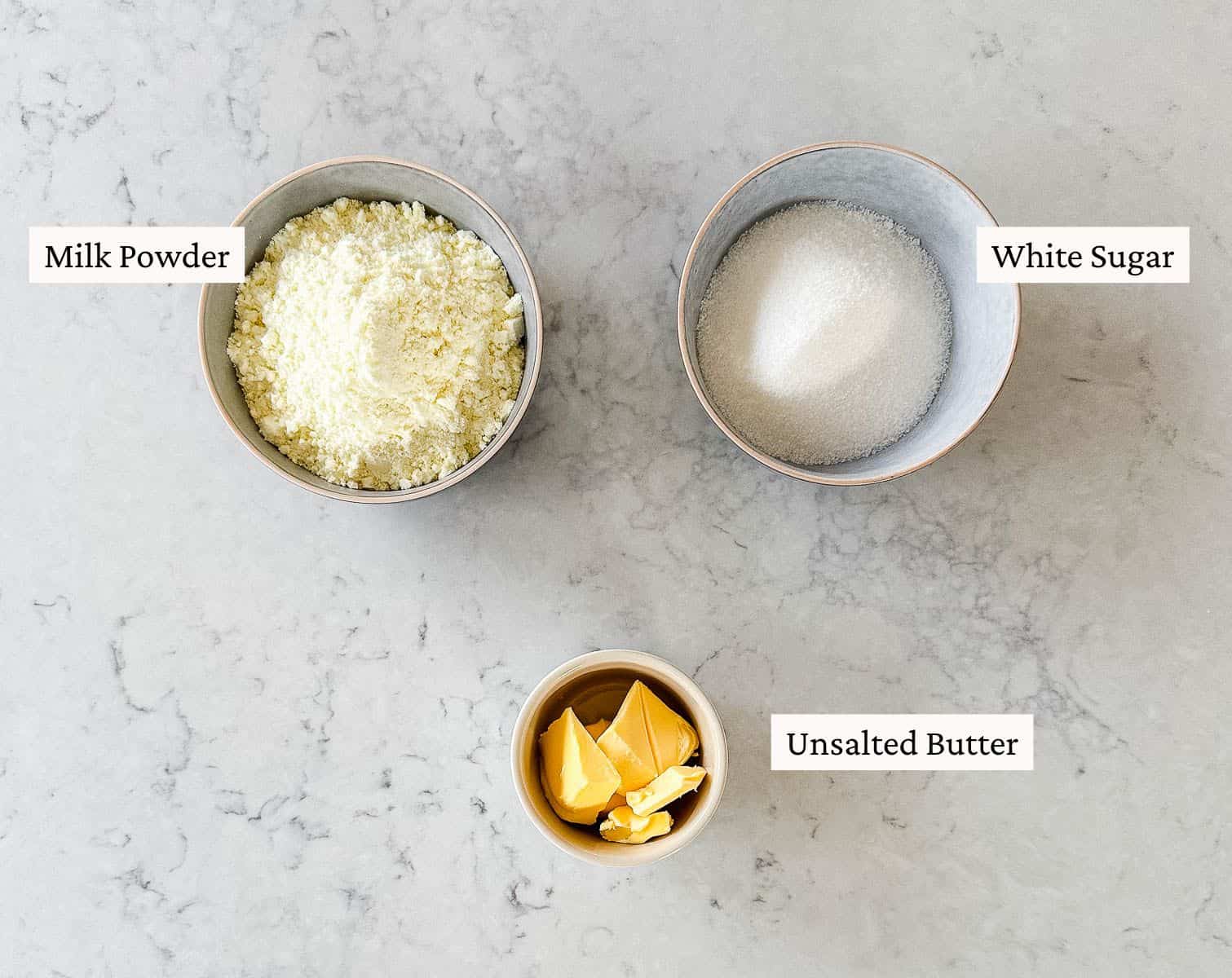 3 bowls filled with milk powder, white sugar and butter, all labelled, on a grey marble bench top.