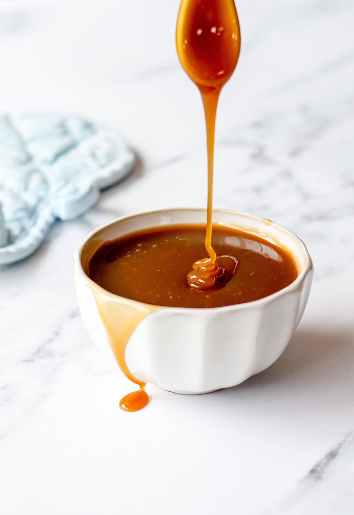 A white bowl filled with caramel sauce sits on a marble surface, with caramel dripping from a spoon above into the bowl and a small spill on the side.