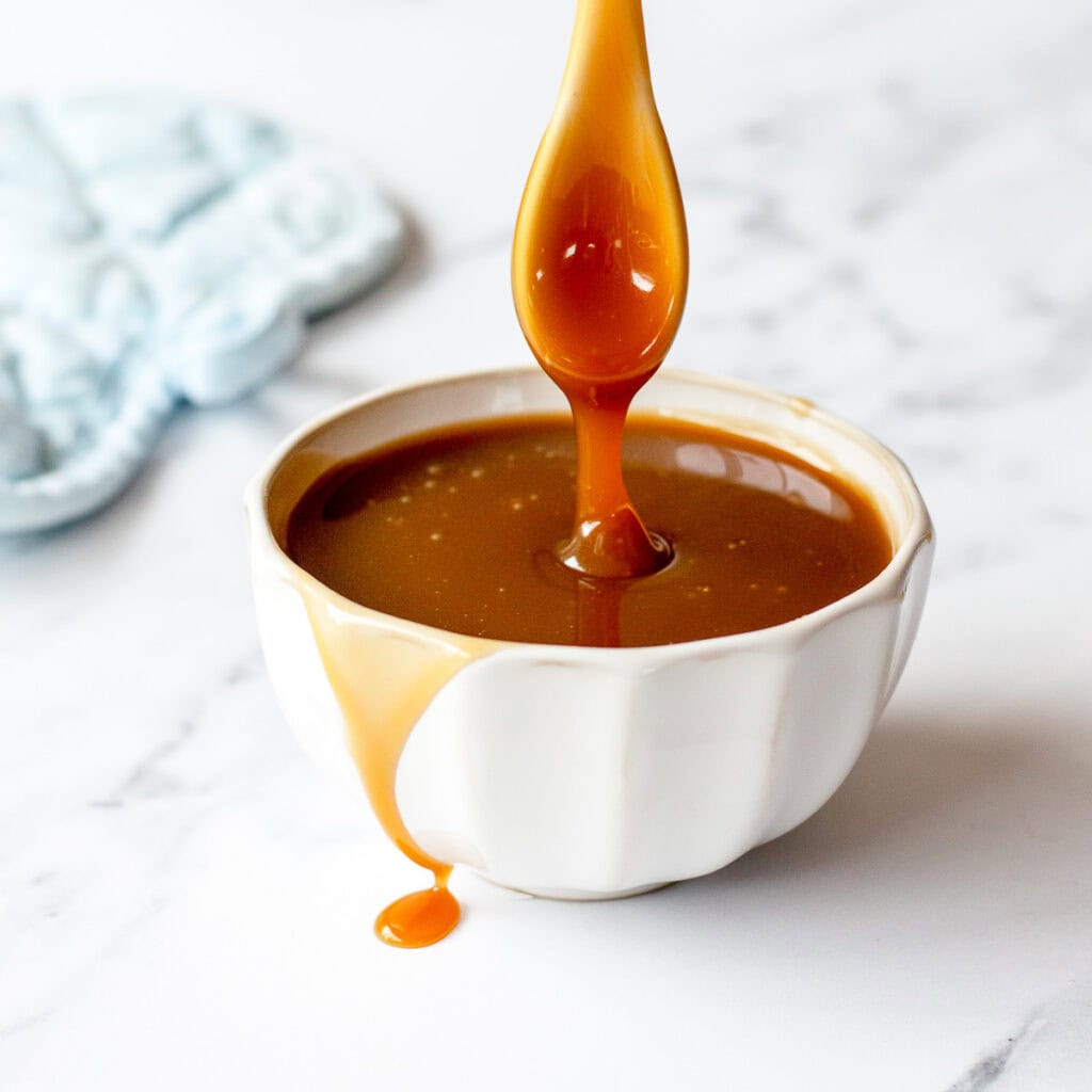 Caramel sauce in a white bowl, dripping from a spoon, on a marble benchtop.