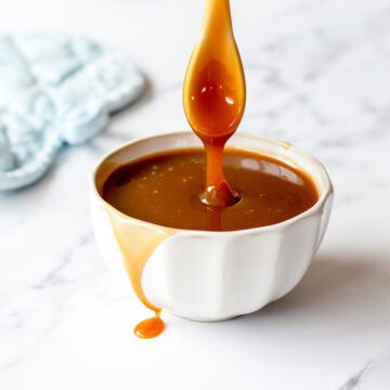 Caramel sauce in a white bowl, dripping from a spoon, on a marble benchtop.