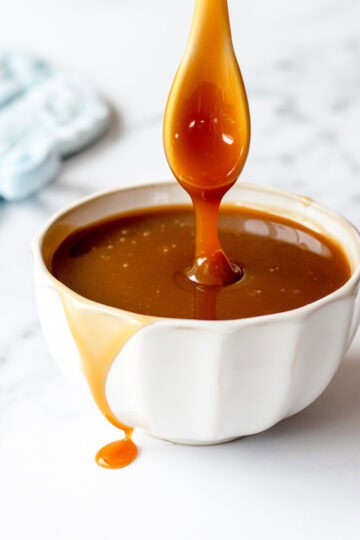 Caramel sauce in a white bowl, dripping from a spoon, on a marble benchtop.