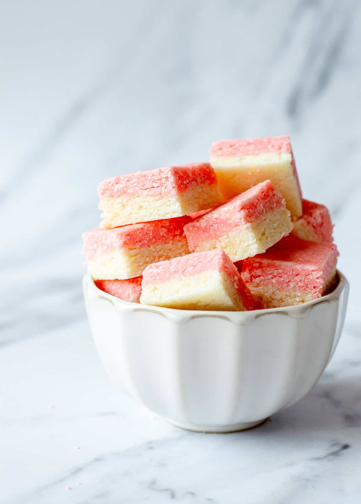 A white bowl filled with pink and white layered coconut squares sits on a marble surface with a blurred background.