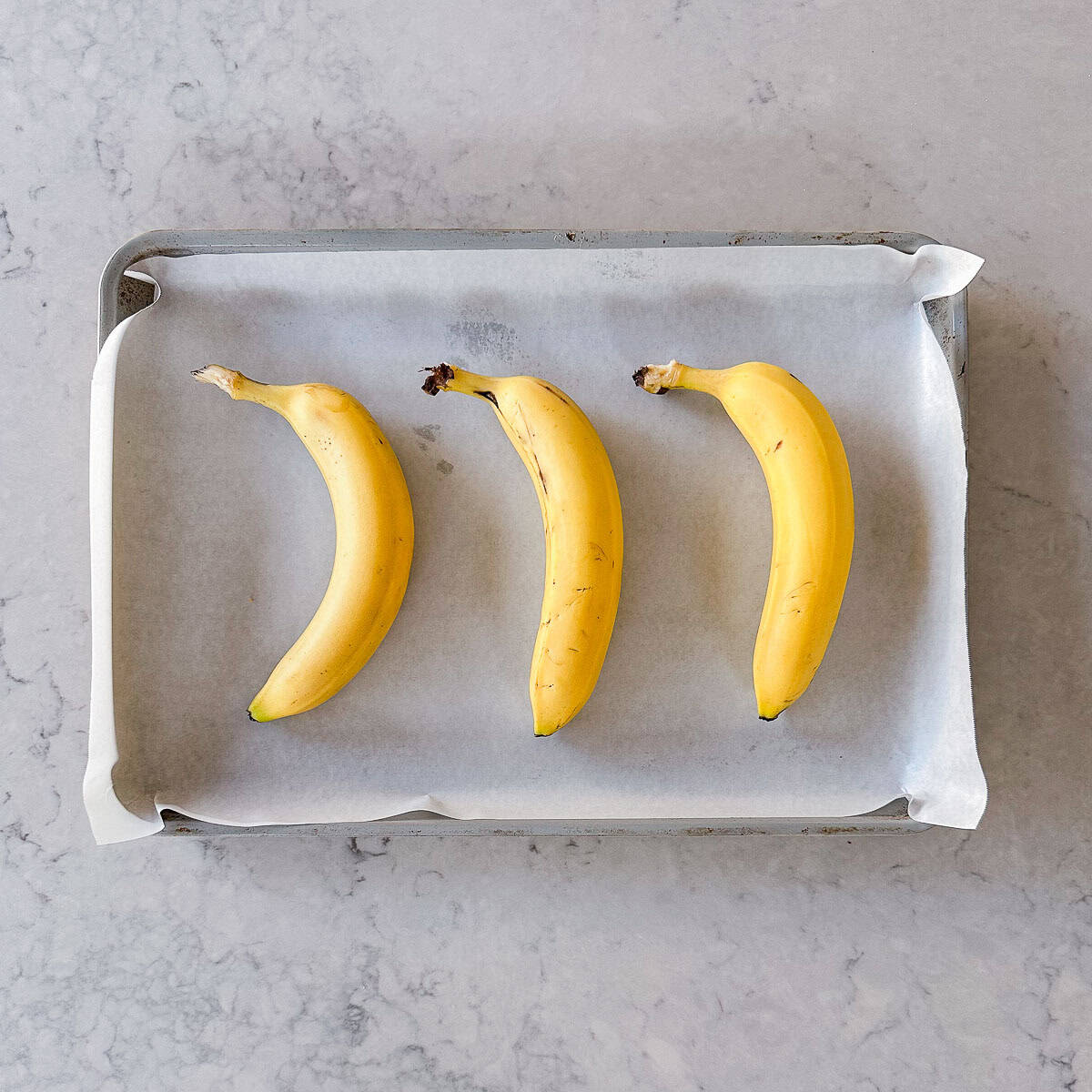Three yellow bananas placed in a row on a baking tray lined with baking parchment, set on a light grey marble worktop.