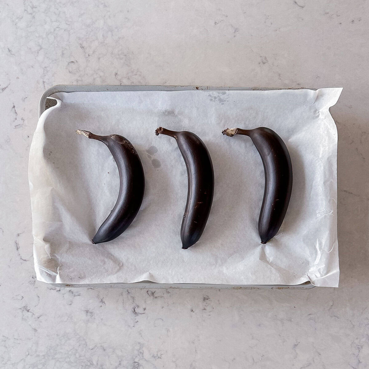 Three whole overripe bananas with black skins are placed in a row on a parchment-lined baking tray, set on a light-coloured marble worktop.