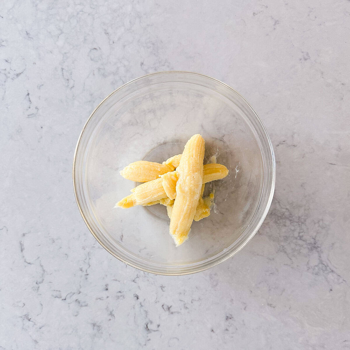 A glass bowl containing peeled bananas cut into pieces, placed on a light grey marble surface.