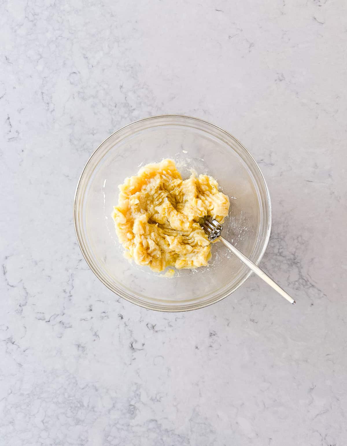 A glass bowl with mashed bananas and a fork inside, placed on a light marble worktop.