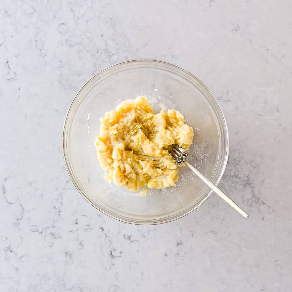 A glass bowl with mashed bananas and a fork inside, placed on a light marble worktop.