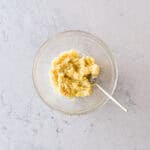 A glass bowl with mashed bananas and a fork inside, placed on a light marble worktop.