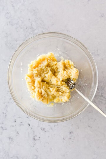 A glass bowl with mashed bananas and a fork inside, placed on a light marble worktop.