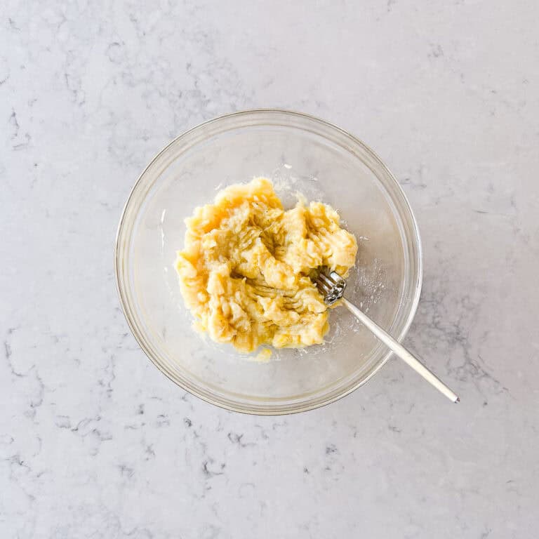 A glass bowl with mashed bananas and a fork inside, placed on a light marble worktop.