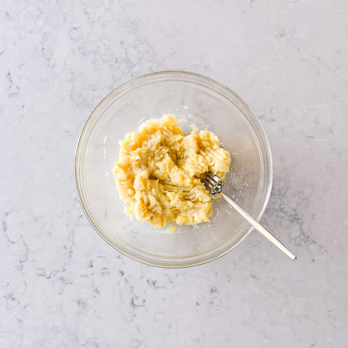 A glass bowl with mashed bananas and a fork inside, placed on a light marble worktop.