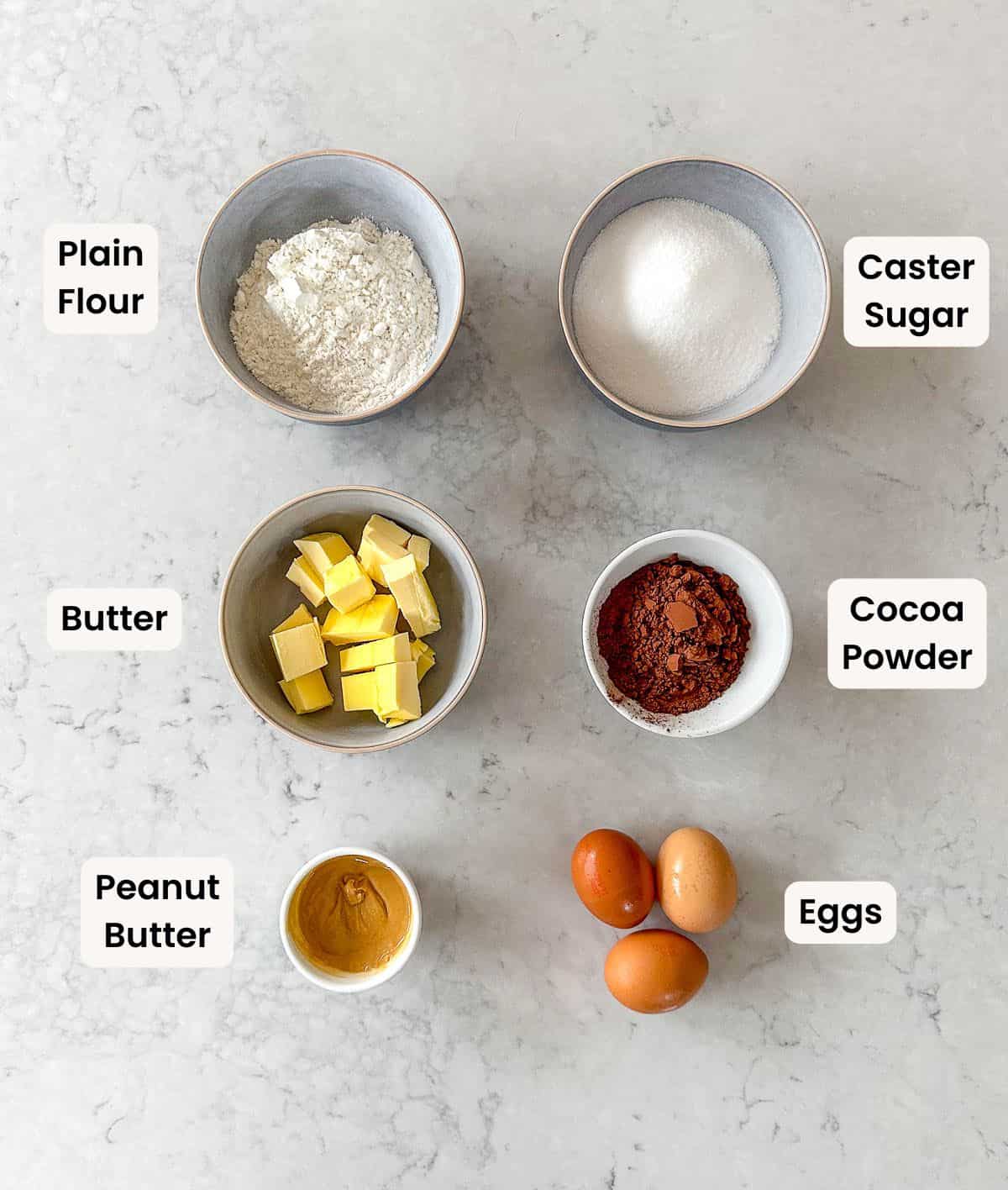 Six bowls and containers on a light surface hold plain flour, caster sugar, cubed butter, cocoa powder, peanut butter, and three eggs, each labelled with its ingredient name.