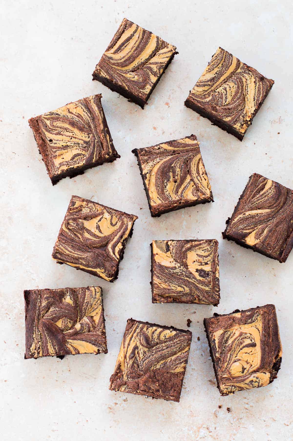 Nine chocolate brownies with a light brown, marbled swirled pattern on top are arranged on a light surface. The brownies are cut into even squares and spaced irregularly.