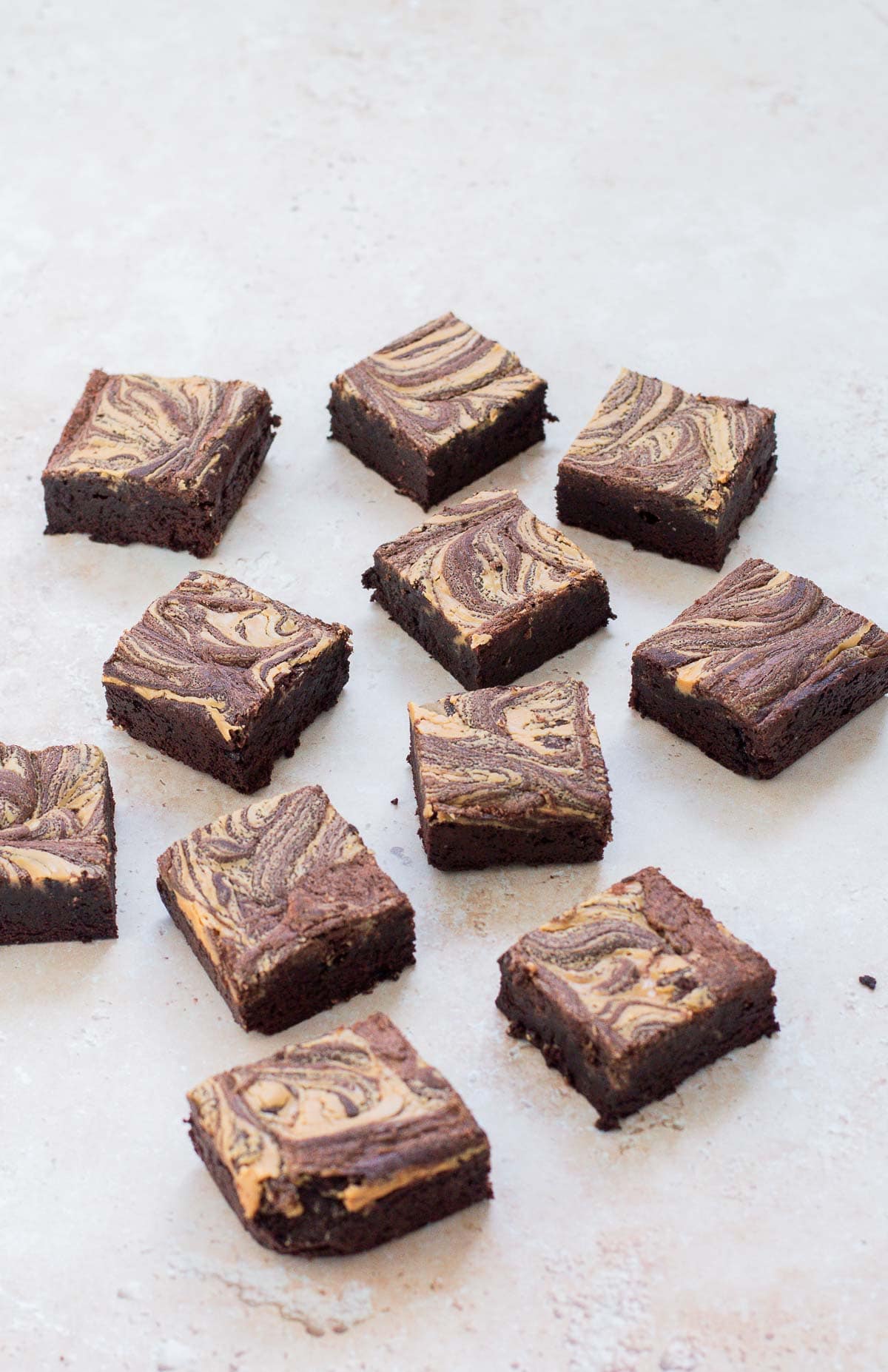 Twelve square brownies with a marbled chocolate and peanut butter swirl pattern are arranged on a light-coloured surface.