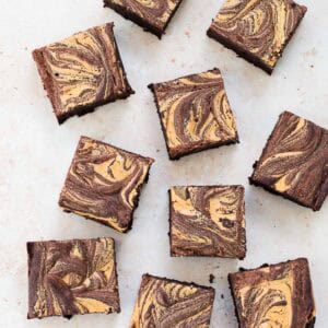 Chocolate brownies with a light brown, marbled swirled pattern on top are arranged on a light surface. The brownies are cut into even squares and spaced irregularly.