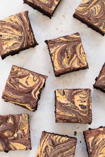 Chocolate brownies with a light brown, marbled swirled pattern on top are arranged on a light surface. The brownies are cut into even squares and spaced irregularly.