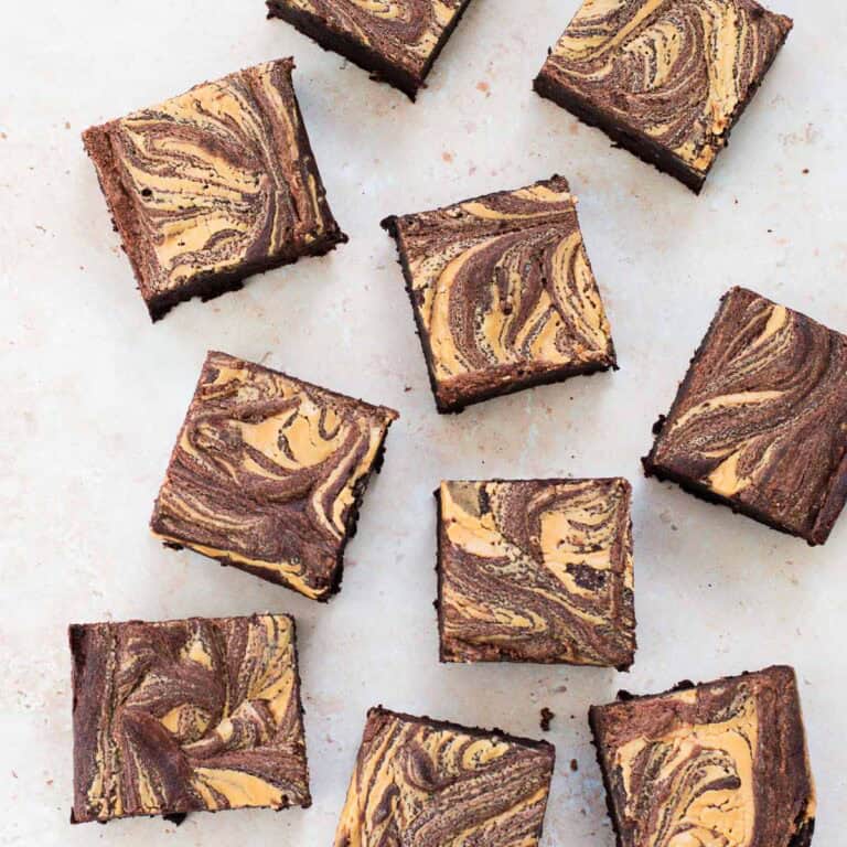 Chocolate brownies with a light brown, marbled swirled pattern on top are arranged on a light surface. The brownies are cut into even squares and spaced irregularly.