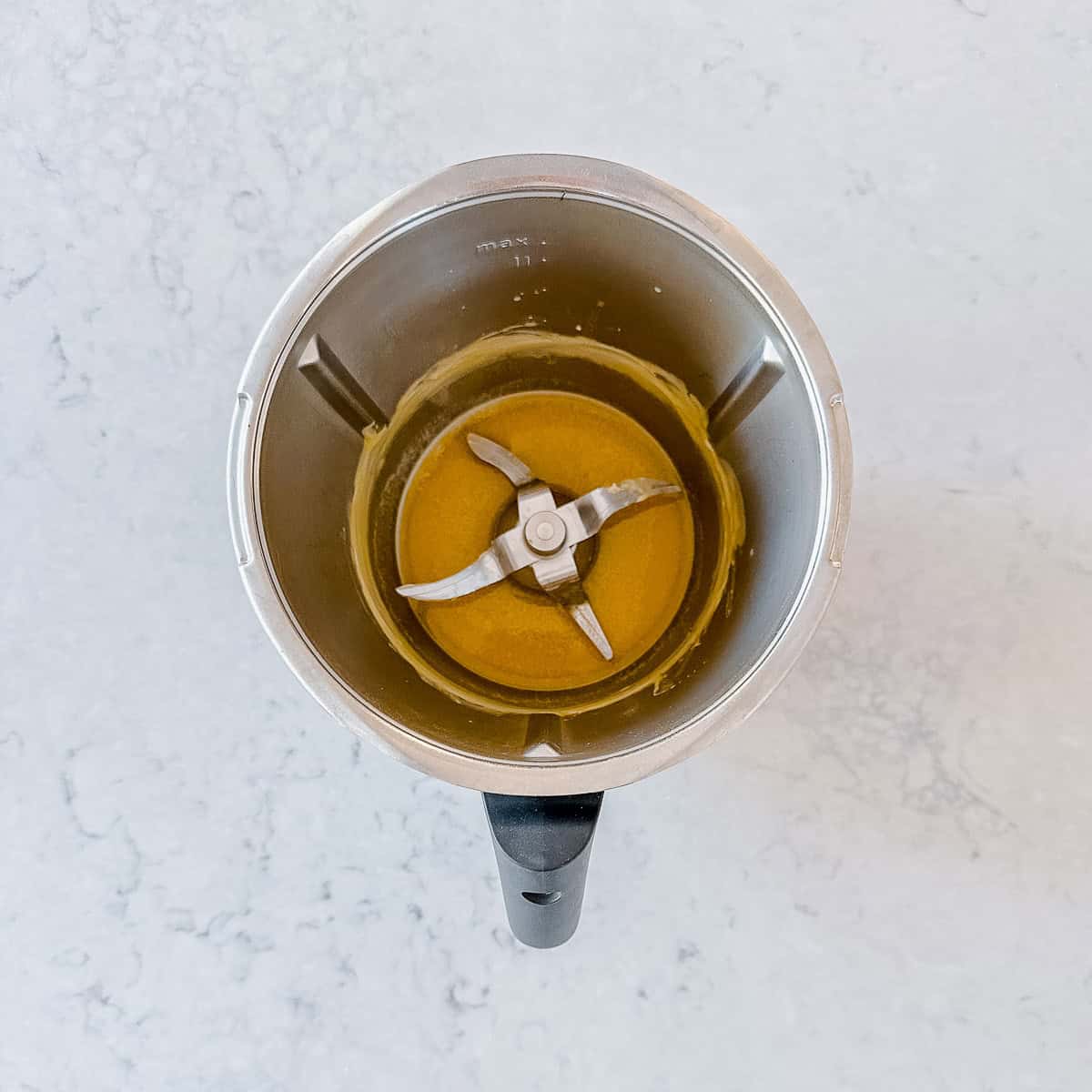 Overhead view of melted butter in the base of a Thermomix bowl, with the blade visible in the centre, on a grey marble benchtop.