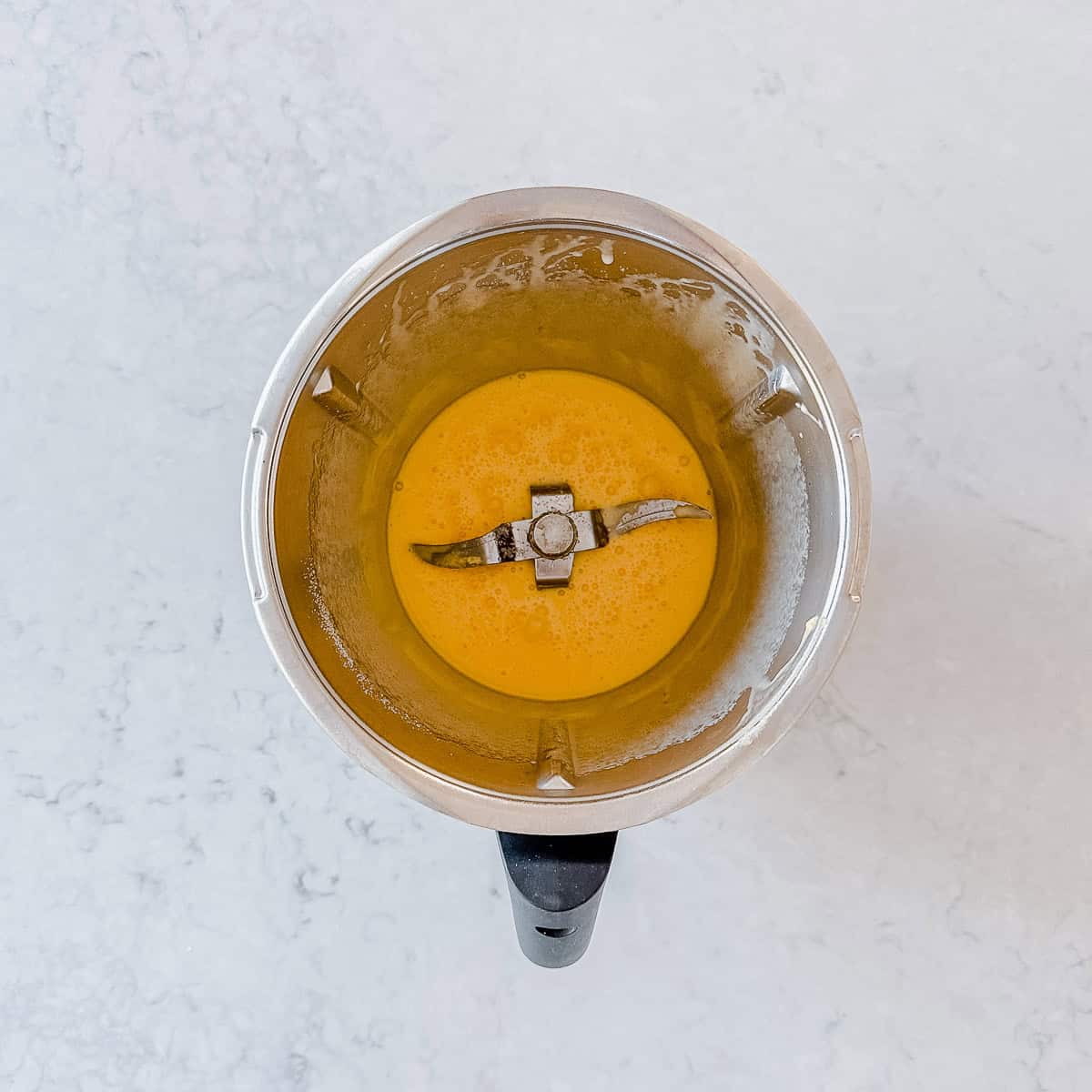 Overhead view of a glossy, bubbly butter, sugar and egg mixture in a Thermomix bowl after mixing.