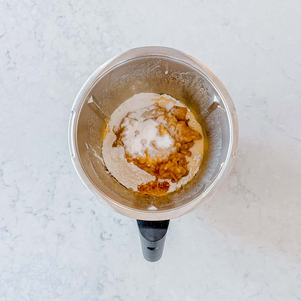 Overhead view of flour, mashed banana and remaining ingredients added to the Thermomix bowl before mixing, showing the unmixed layers.