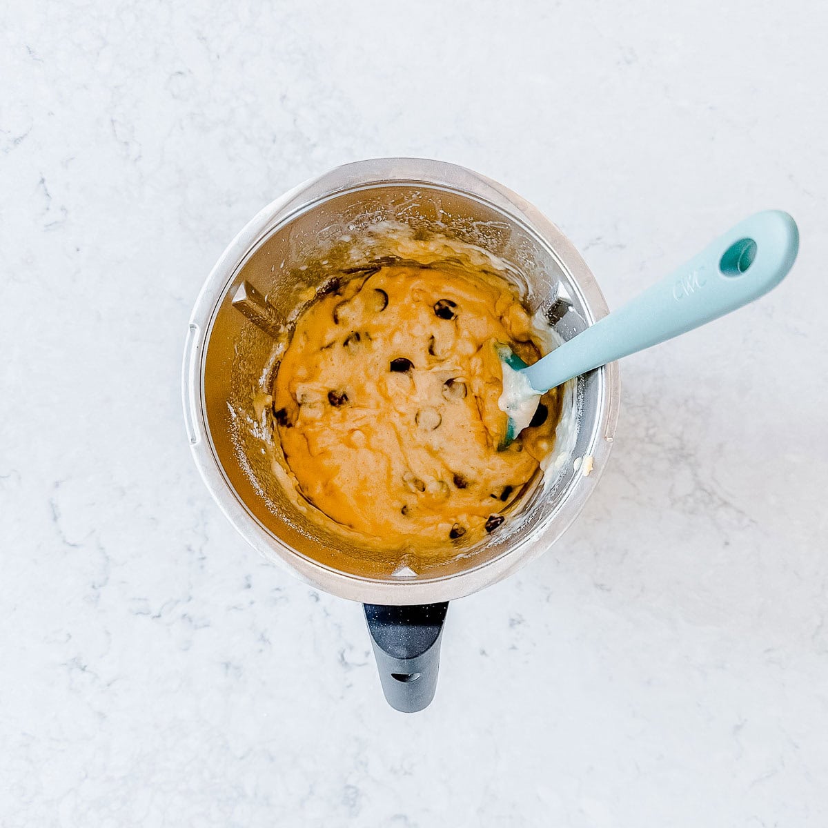 Overhead view of chocolate chips being folded into the banana bread batter with a teal spatula inside a Thermomix bowl.