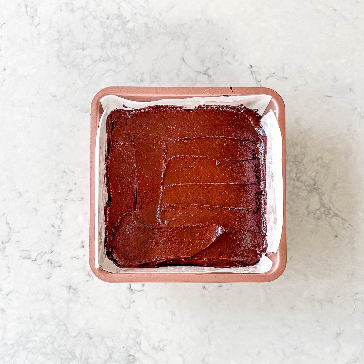 A square baking tin lined with greaseproof paper, filled with chocolate brownie batter, waiting to be baked, on a light marble surface.