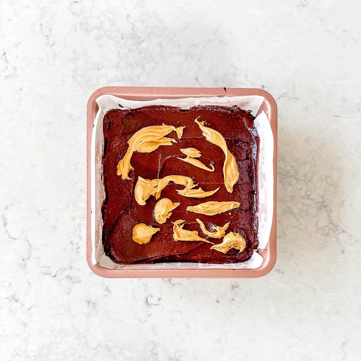 A square tin of chocolate brownies topped with swirls of peanut butter, sitting on a white marble surface. The brownies are still in the tin, lined with baking parchment.