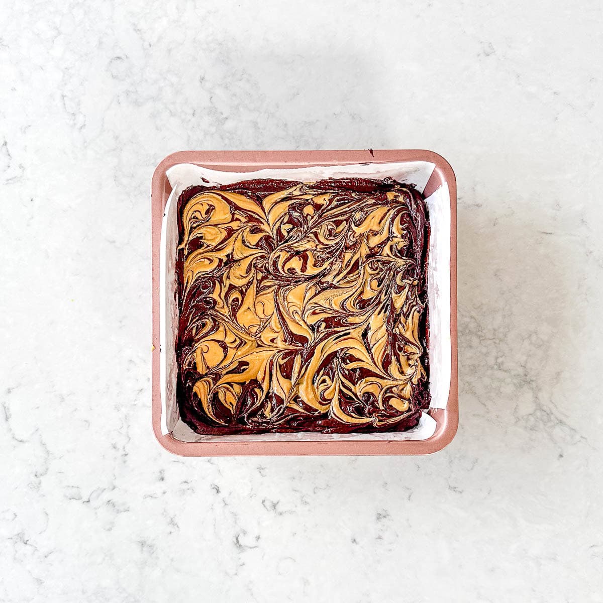 A square tin of brownies with a marbled swirl of chocolate and peanut butter on top, set on a light marble surface.