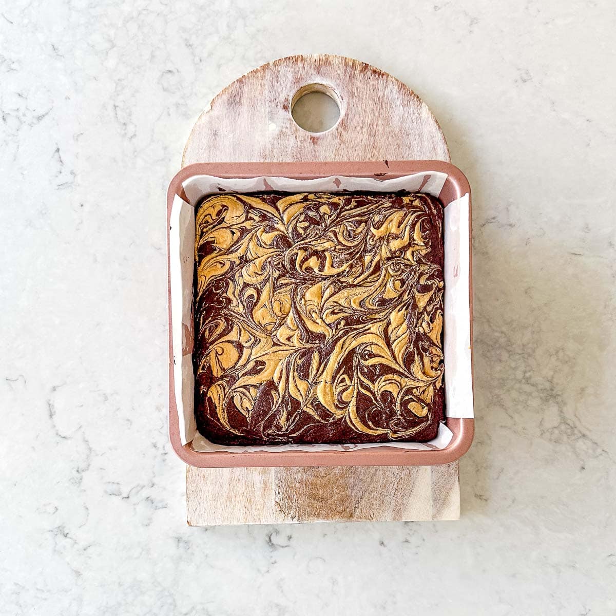 A square tin of brownies with a marbled chocolate and peanut butter pattern on top sits on a small wooden chopping board on a light marble worktop.