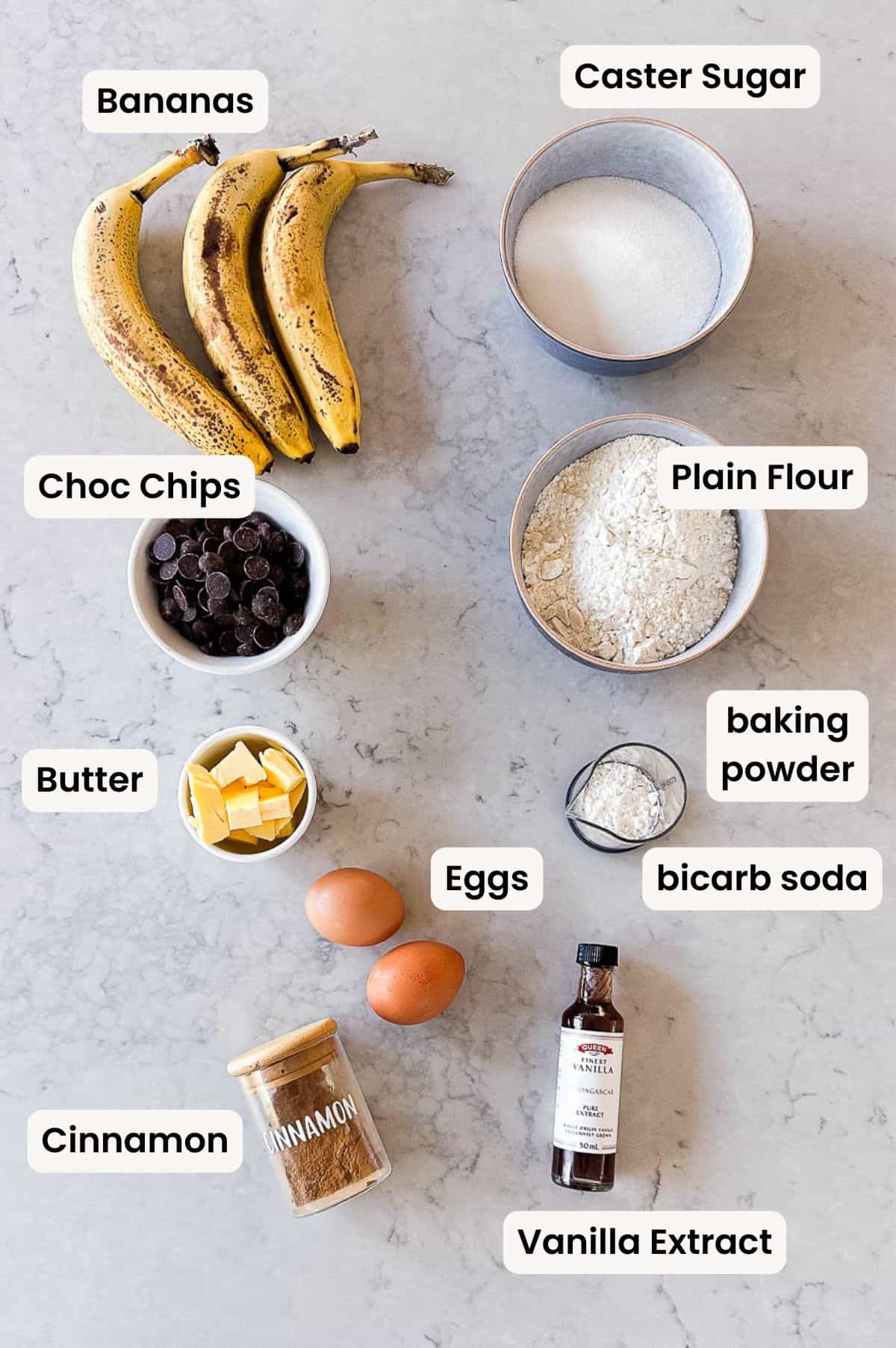 Overhead flat lay of the ingredients needed to make Thermomix Chocolate Chip Banana Bread, including ripe bananas, caster sugar, plain flour, chocolate chips, butter, eggs, baking powder, bicarbonate of soda, cinnamon and vanilla extract, each labelled on a marble surface.