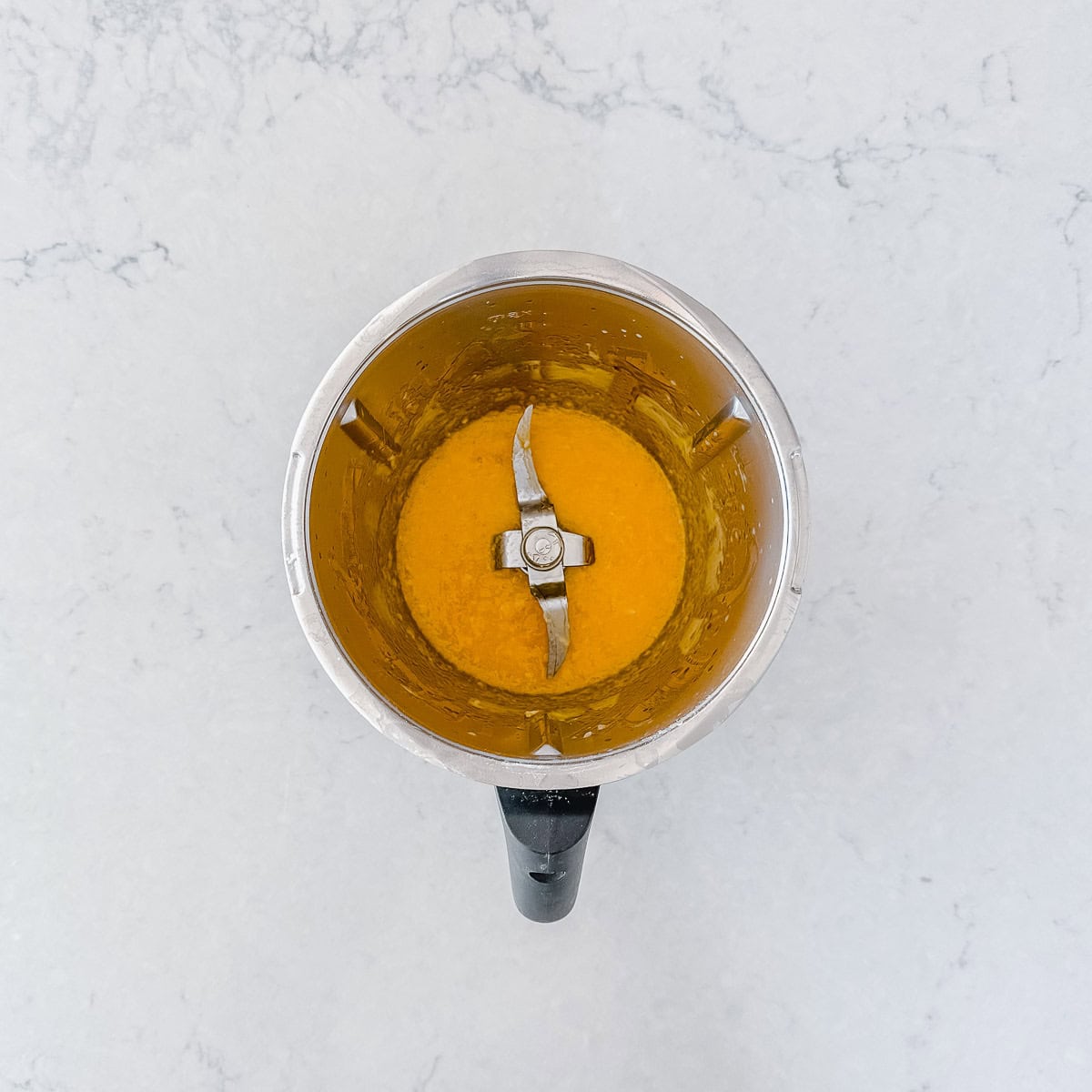 A top-down view of a Thermomix jug with banana purée inside, placed on a light marble worktop.