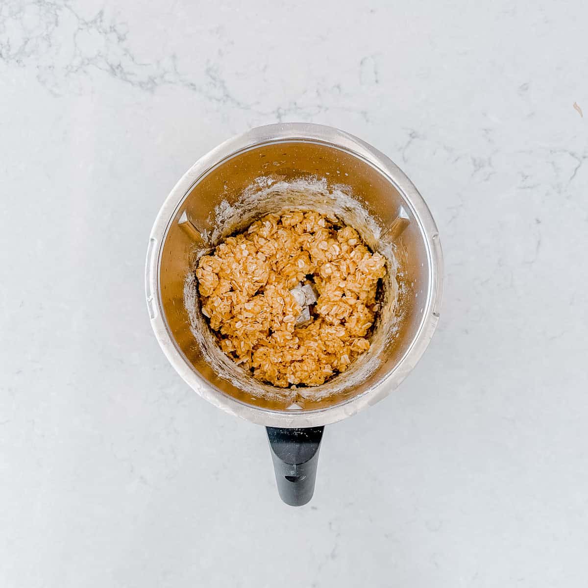A Thermomix jug, containing a mixture of oats and batter, sits on a light marble surface.